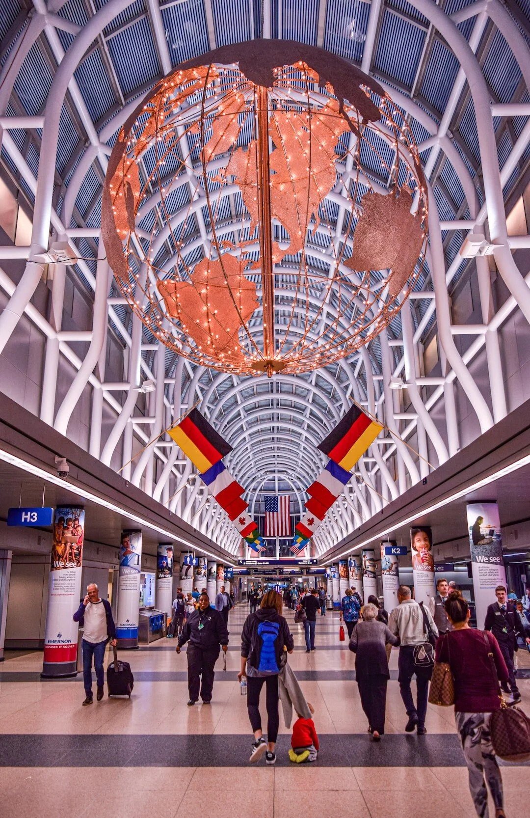 Inside an airport terminal with a large illuminated globe art piece hanging from the ceiling, decorated with world map outlines and lights. Flags of Germany, Canada, France, and other countries line the corridor. Passengers walk through the spacious, modern space.