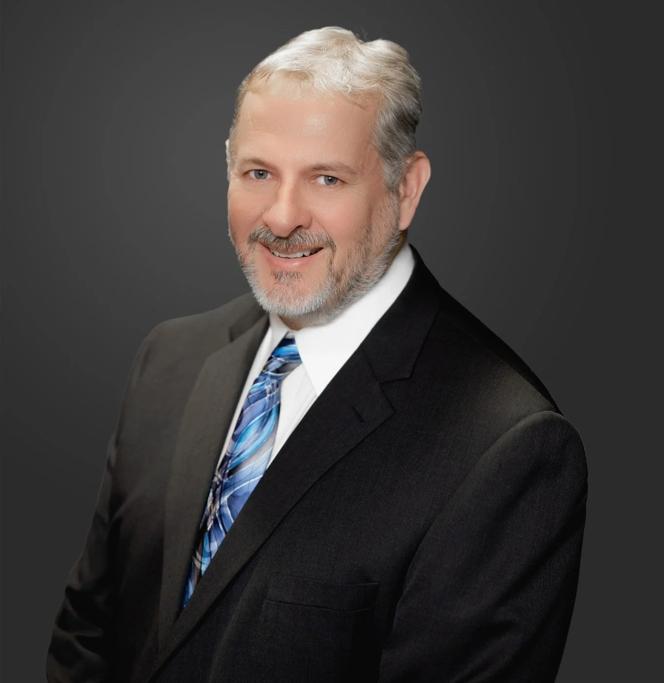 A professional headshot of a smiling middle-aged man with gray hair and beard, wearing a black suit, white shirt, and blue patterned tie, against a dark gray background.