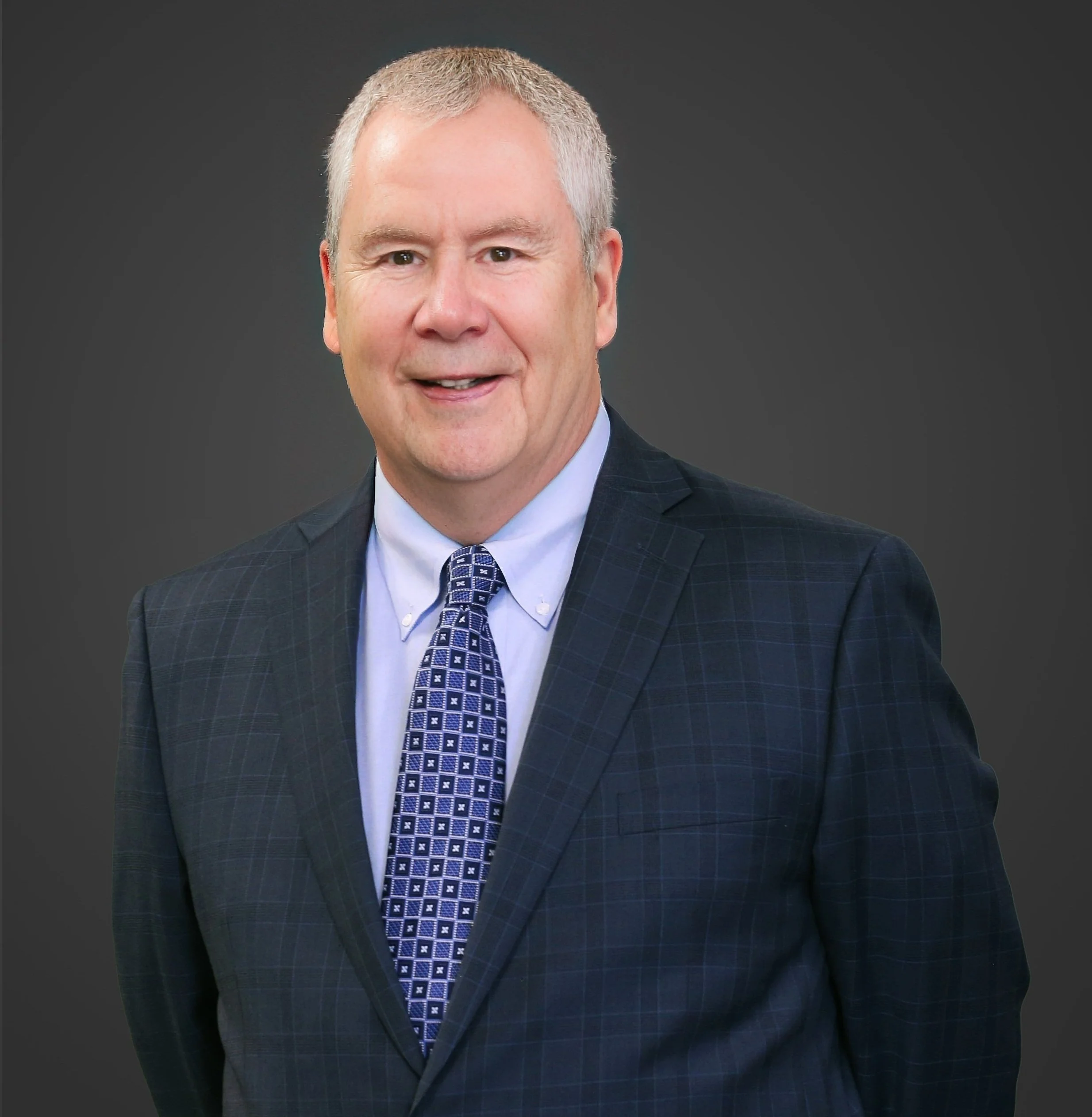 A middle-aged man with short gray hair, wearing a dark blue checkered suit, a light blue shirt, and a blue patterned tie, against a dark background.