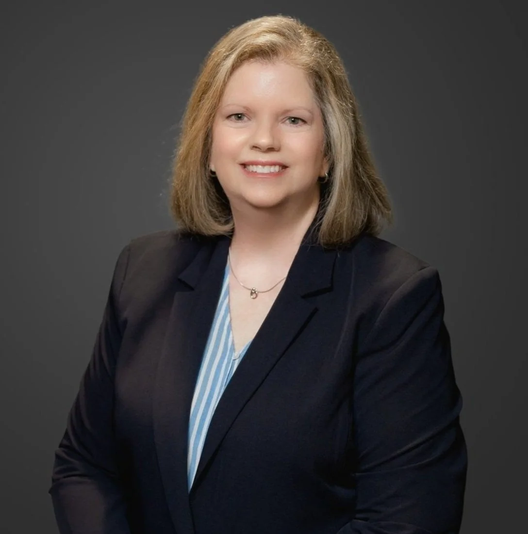 Professional woman with shoulder-length blonde hair, smiling, wearing a dark blazer and light blue blouse against a dark gray background.