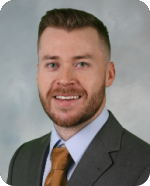 Professional headshot of a man in a suit and tie, smiling, with short dark hair and a beard