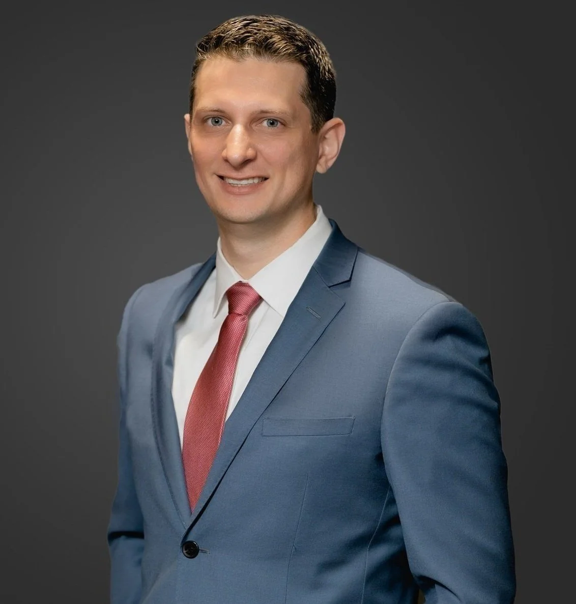 Professional portrait of a smiling man in a blue suit and pink tie against a dark background.