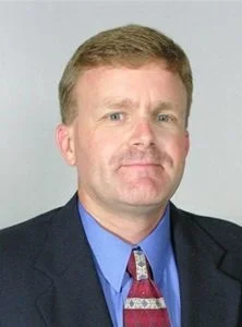 Portrait of a middle-aged man with light brown hair, blue eyes, wearing a dark suit, blue shirt, and patterned tie, against a plain background.