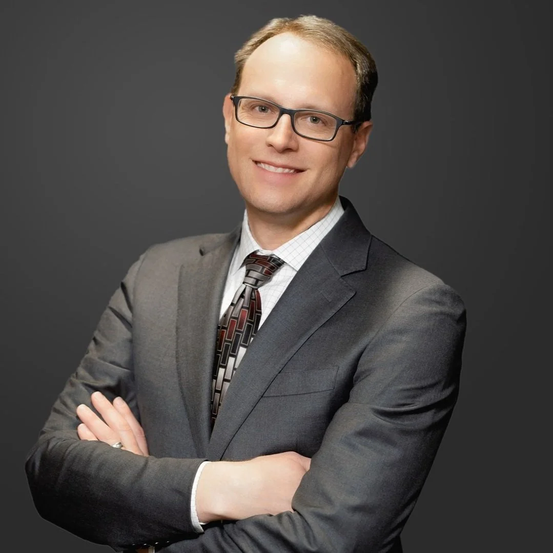 A man in a gray suit, checkered shirt, and patterned tie, wearing glasses, standing with arms crossed and smiling against a gray background.