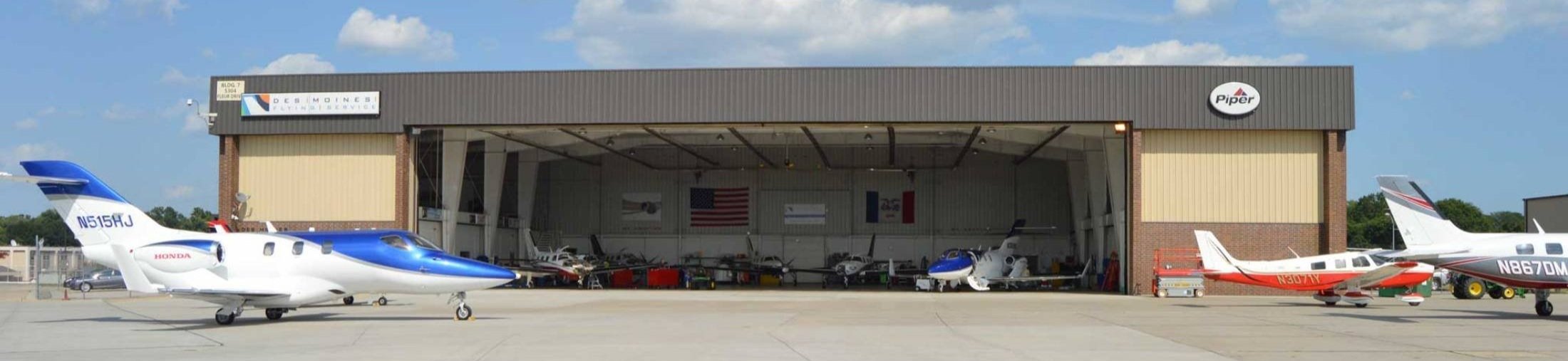 Aircraft hangar with several small planes parked outside, including a blue and white jet on the left and a red and white plane on the right, under a partly cloudy sky.