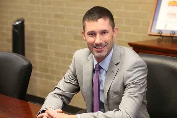 A man in a gray suit and purple tie sitting at a conference table, smiling at the camera, with a brick wall and a framed picture in the background.