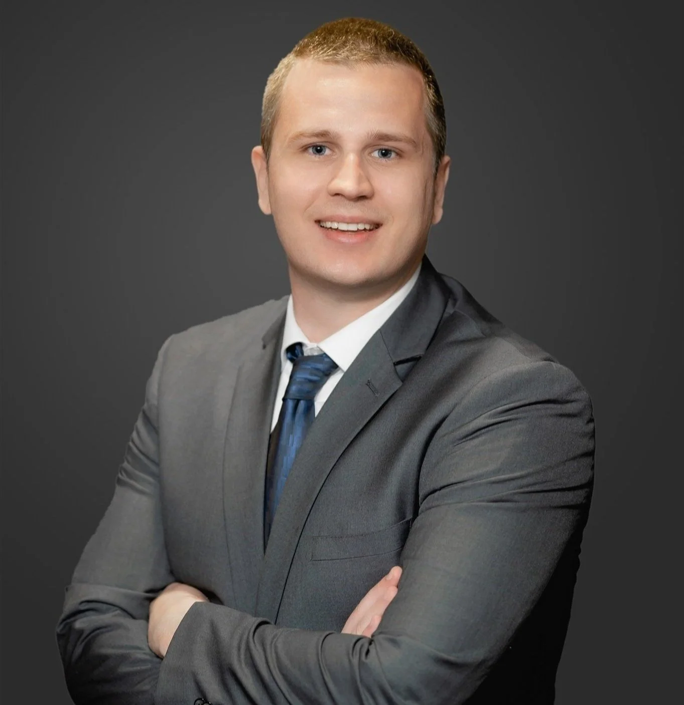 A young man with short blonde hair wearing a light grey suit, white shirt, and dark tie, posing with arms crossed against a dark grey background.