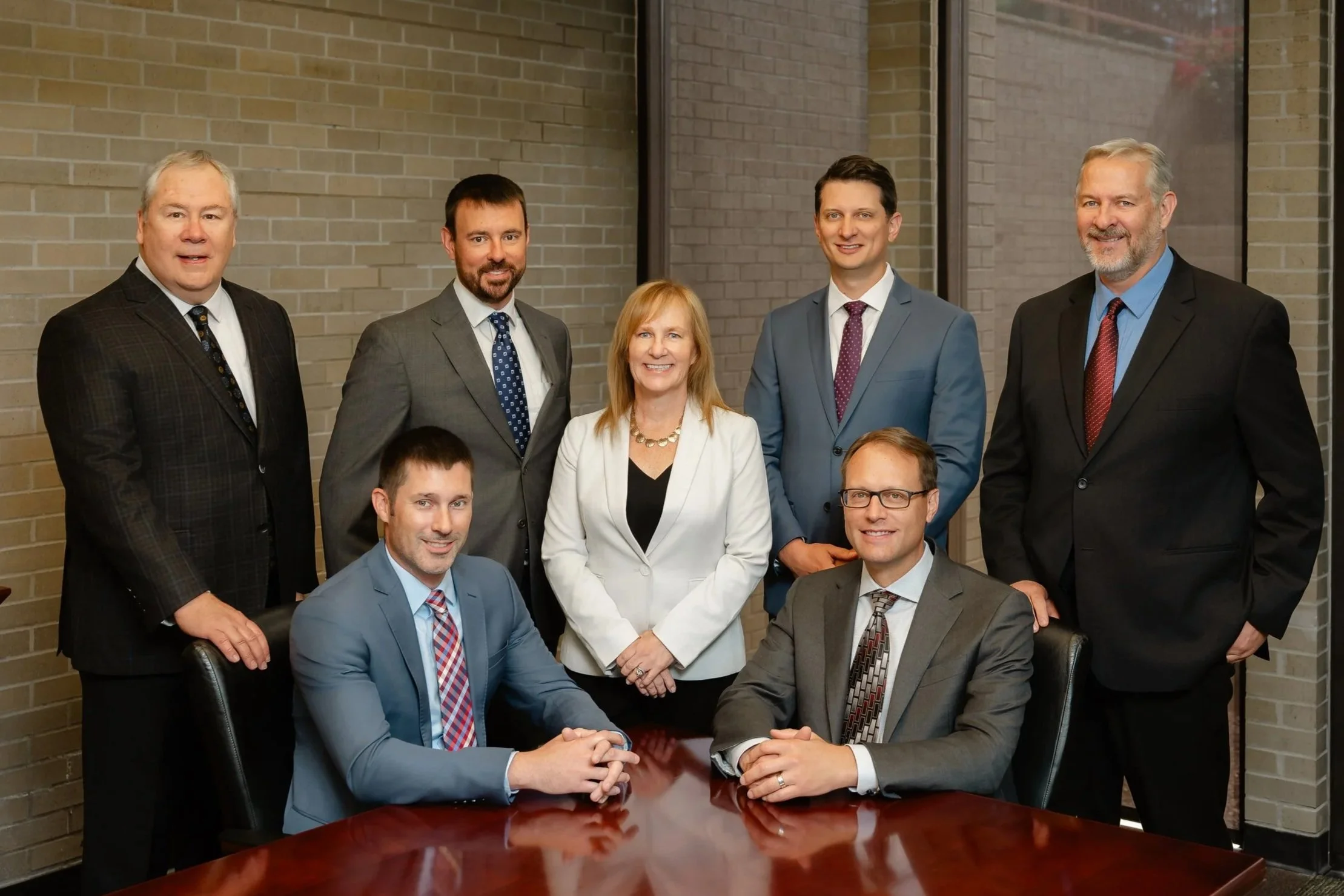 Group photo of nine professionally dressed individuals, five men and one woman, posing in front of a brick wall. Some are standing, others seated around a wooden conference table, smiling at the camera.