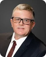 A man wearing glasses, a dark suit, white shirt, and red tie, with short light-colored hair, against a gray background.