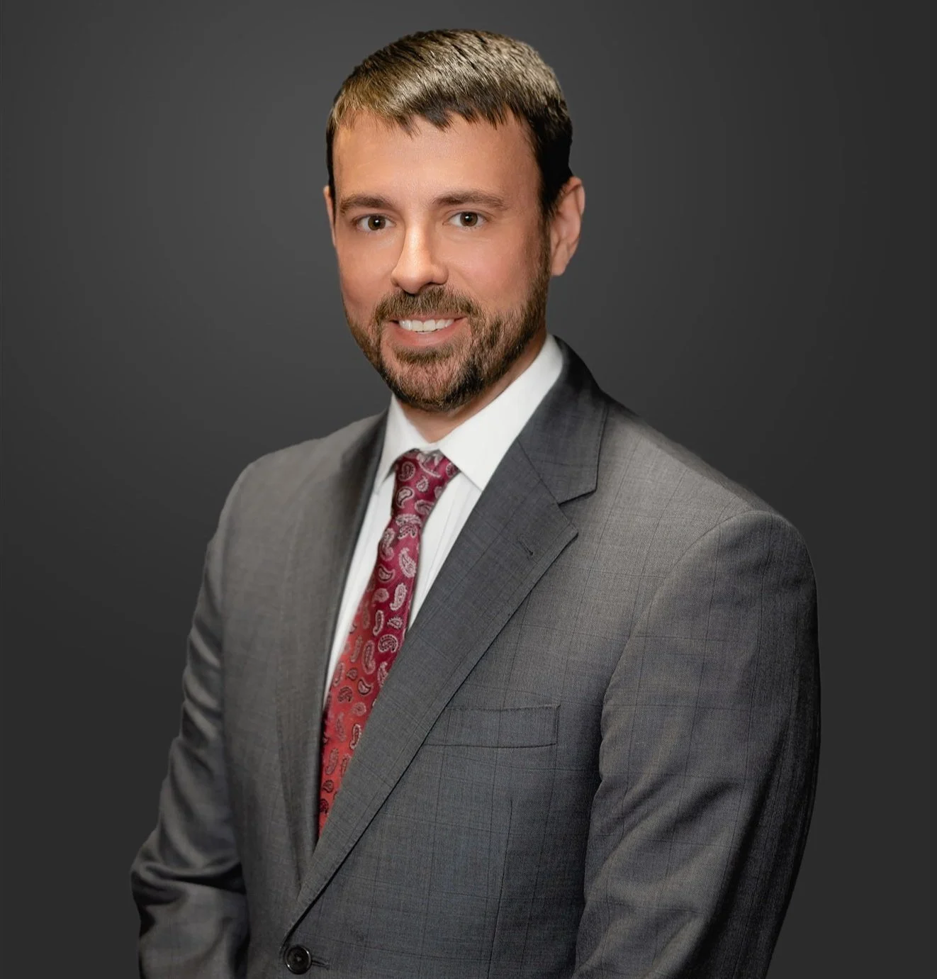 A professional man with a beard and brown hair wearing a gray suit, white shirt, and red patterned tie, smiling against a dark gray background.