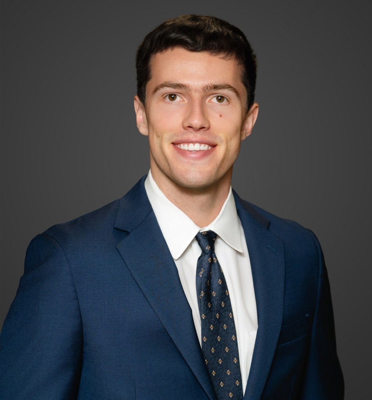 A young man with short dark hair, wearing a navy blue suit, white shirt, and a patterned tie, smiling against a dark gray background.