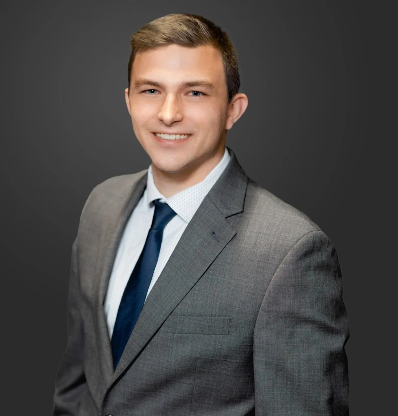 A young man in a gray suit, white shirt, and navy tie, smiling against a dark gray background.