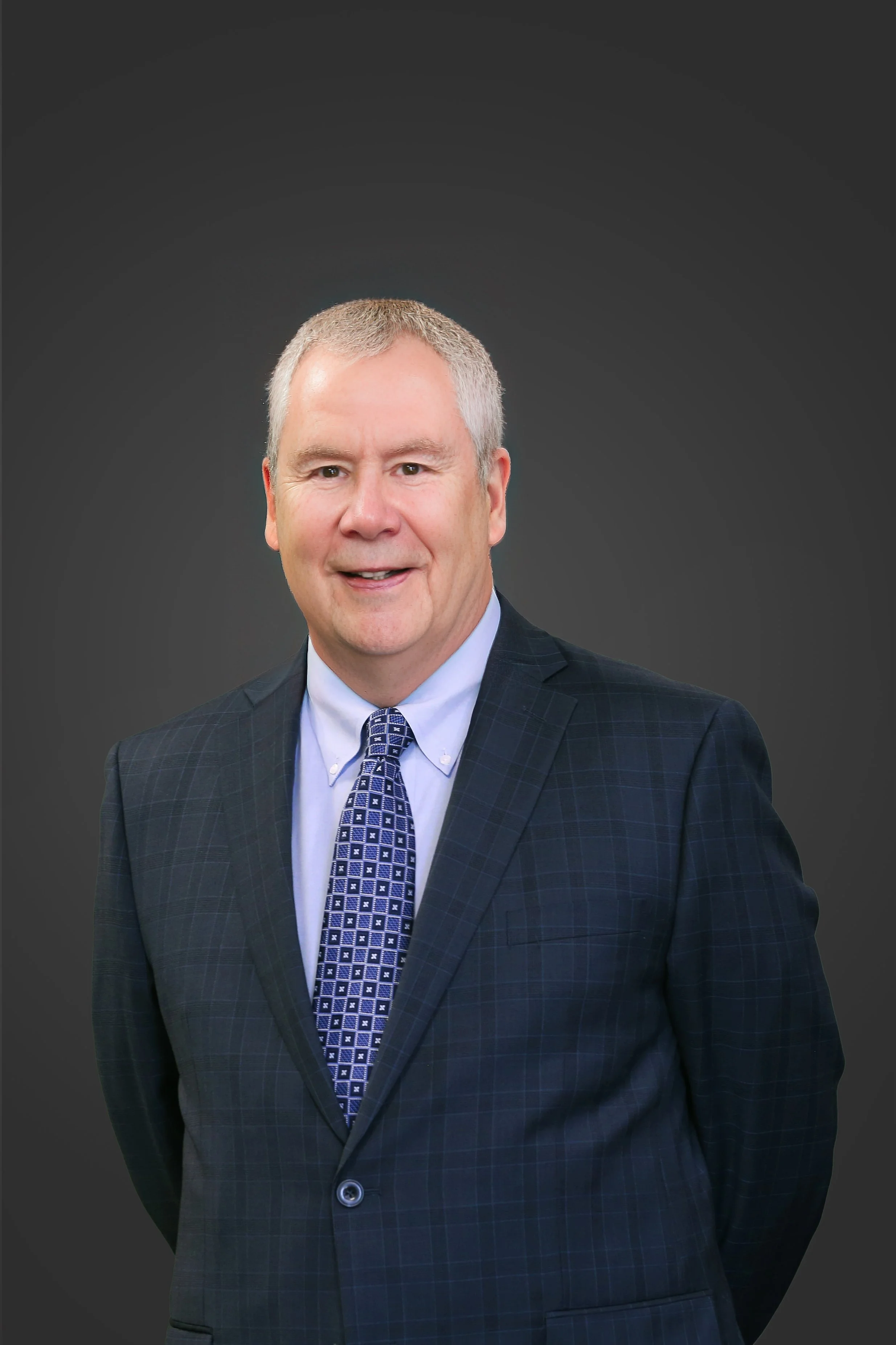 A man with short gray hair dressed in a dark plaid suit, light blue shirt, and a patterned blue tie, smiling against a dark background.