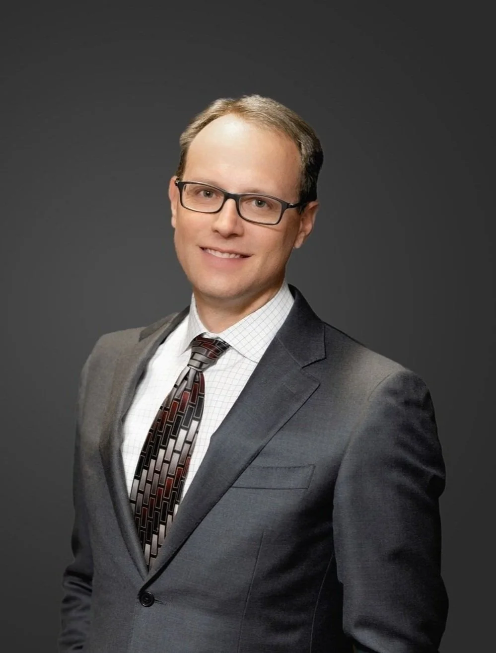 Portrait of a man in a dark gray suit, white dress shirt, and patterned tie, wearing glasses, smiling against a plain dark background.