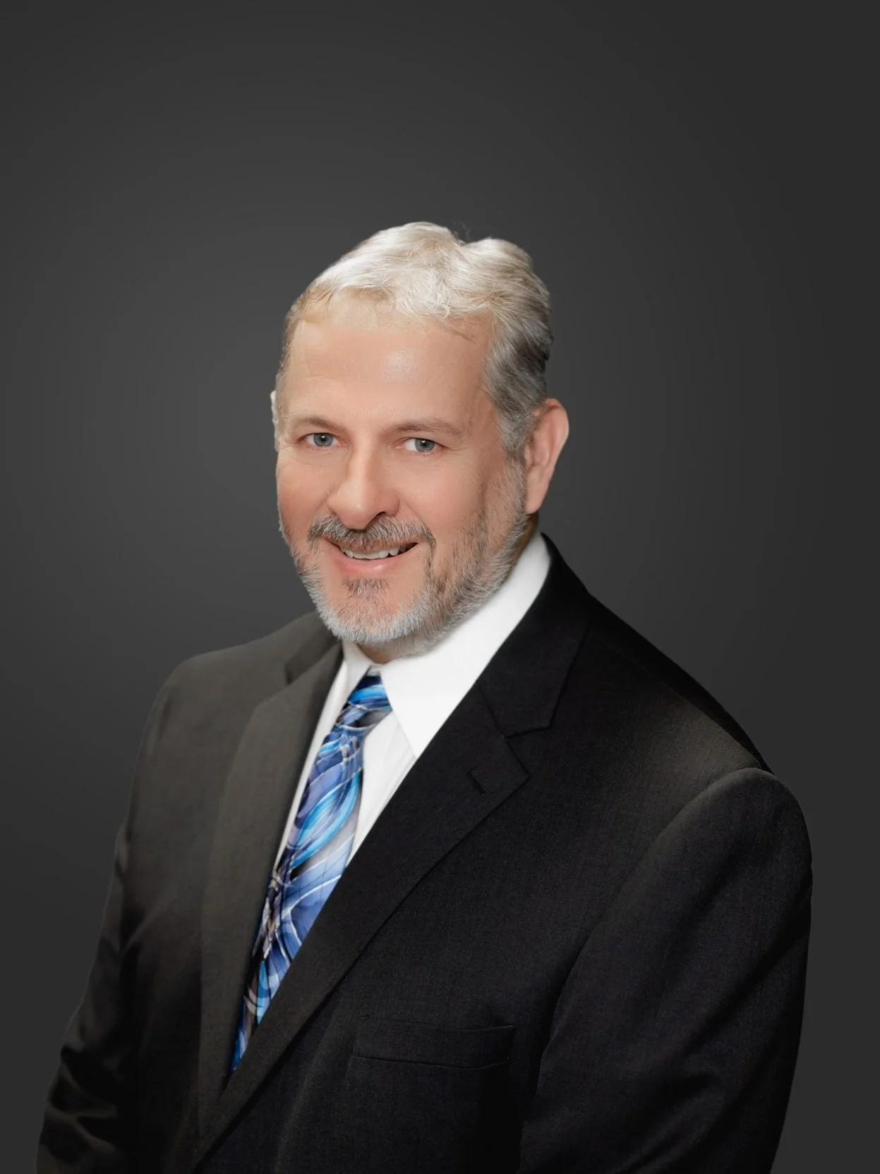 A professional headshot of a middle-aged man with gray hair, beard, and blue eyes, wearing a dark suit, white shirt, and a blue patterned tie, smiling against a dark gray background.