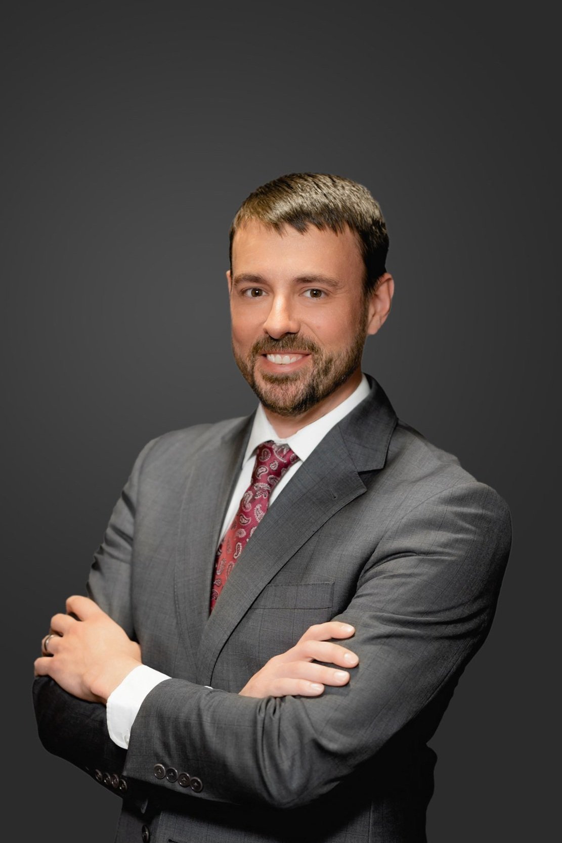 A man in a gray suit with a patterned tie, smiling, with arms crossed, against a dark gray background.