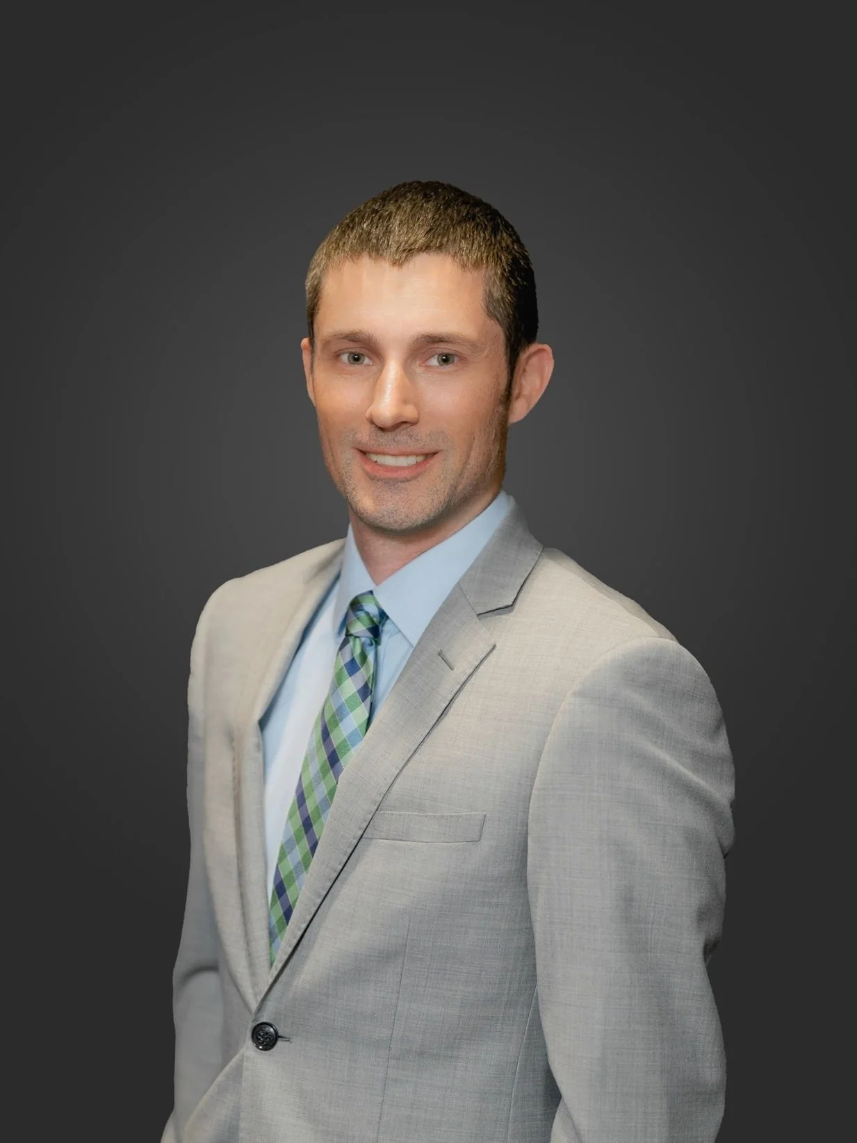 Portrait of a young man with short brown hair, wearing a light gray suit, light blue shirt, and green plaid tie, standing against a dark gray background.