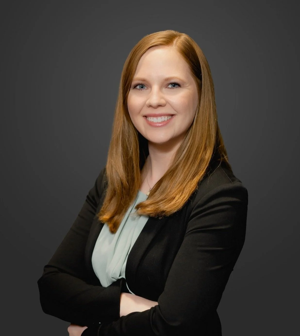 Portrait of a smiling woman with long red hair, wearing a black blazer and a light-colored blouse, against a dark gray background.