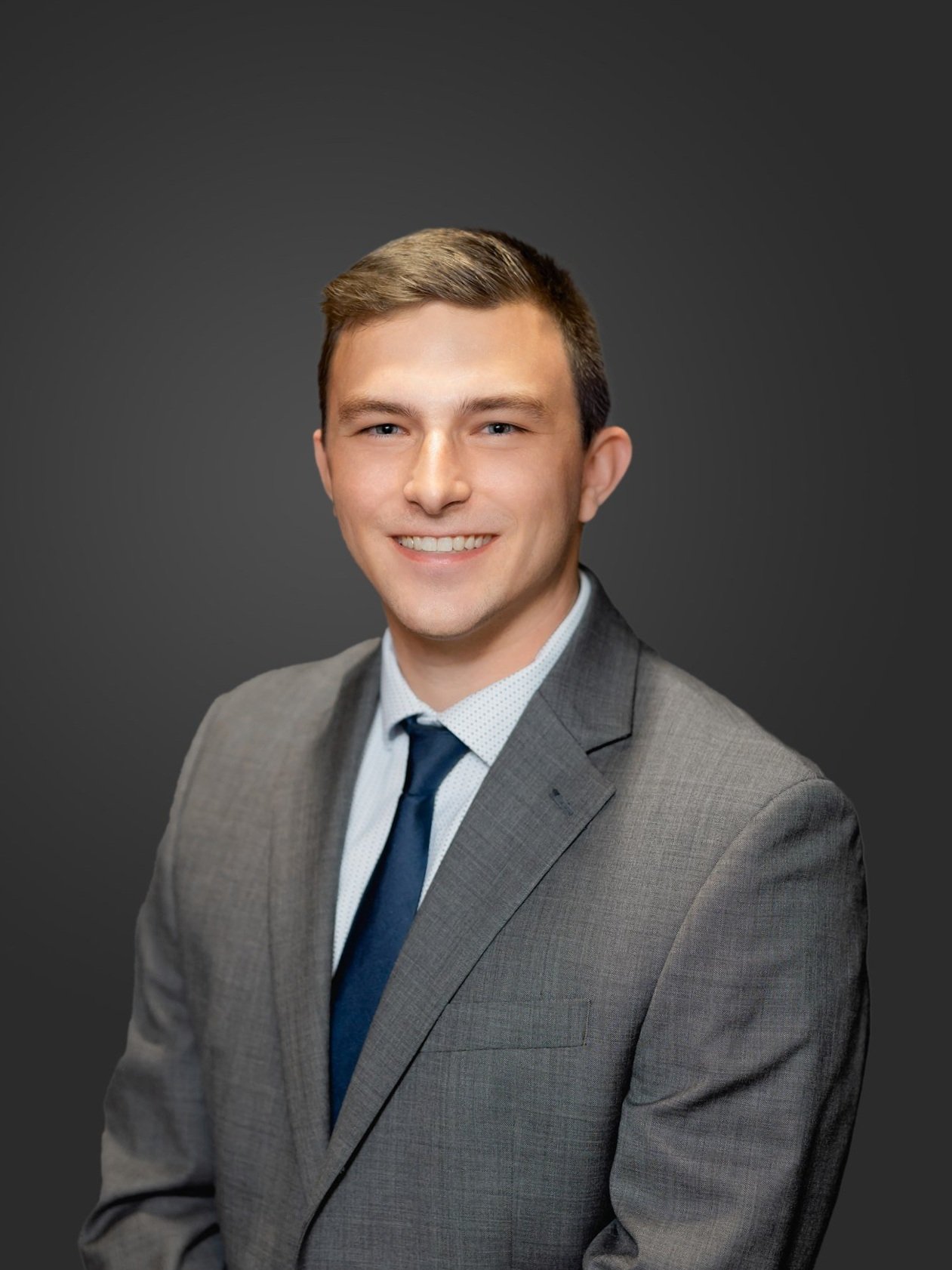 A young man in a gray suit with a white shirt and navy tie, smiling, against a dark gray background.