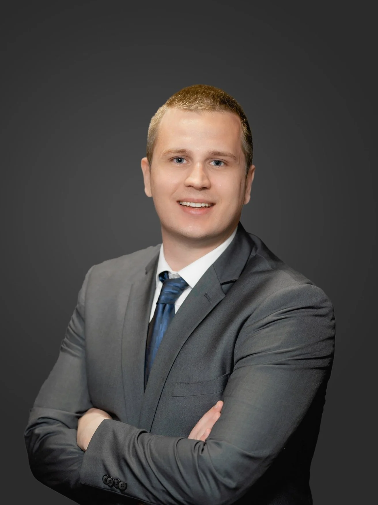 A young man in a gray suit and blue tie posing against a dark background, smiling with arms crossed.