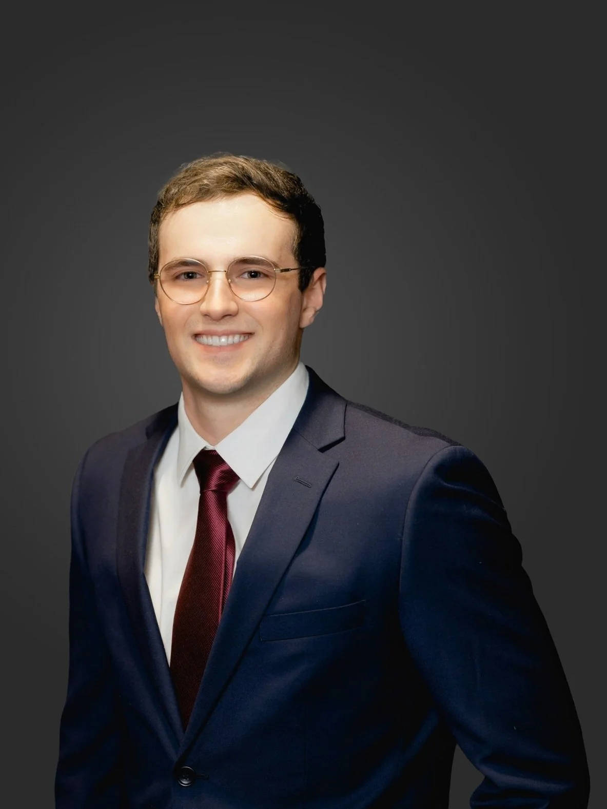 Portrait of a young man in a business suit, wearing glasses, smiling, with dark background.