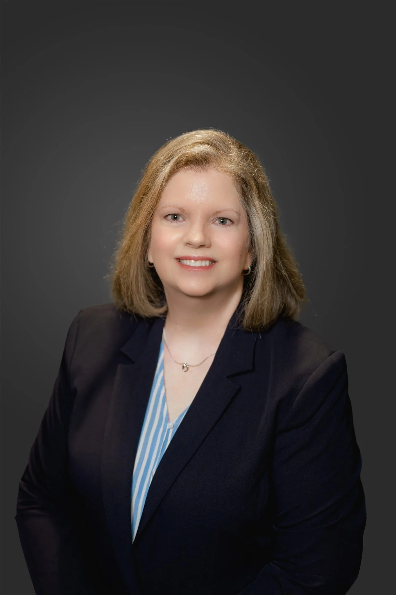 A woman with shoulder-length blonde hair, wearing a dark blazer over a light blue and white striped shirt, smiling against a dark gray background.