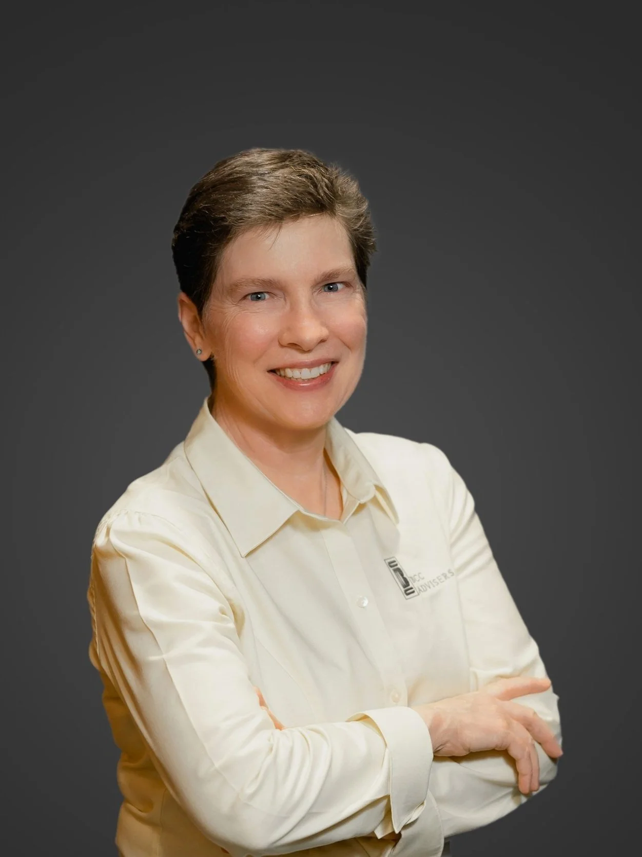 A woman with short brown hair smiling in a cream-colored button-up shirt with a company logo, standing against a dark gray background.