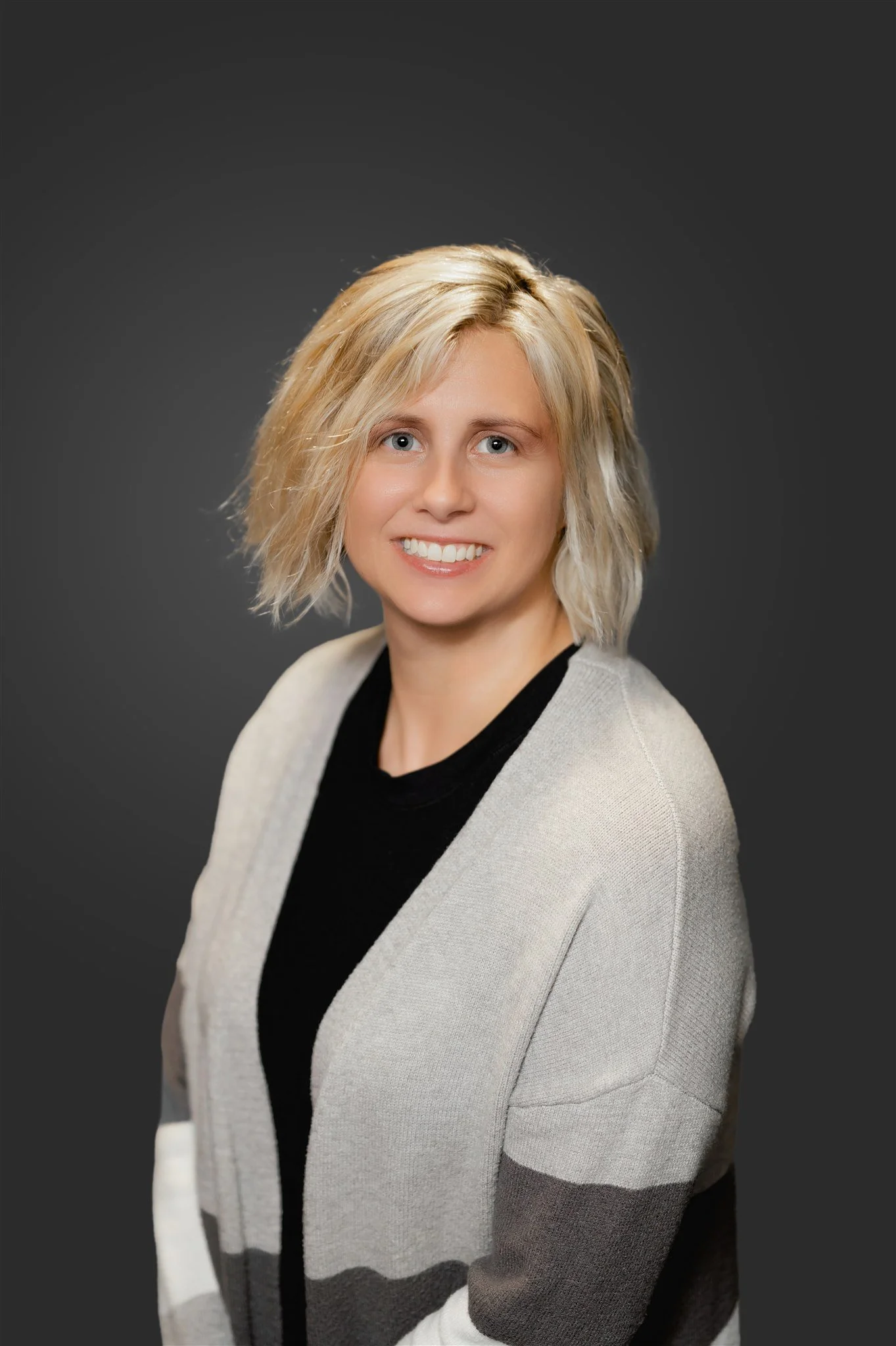 Portrait of a smiling woman with short blonde hair, wearing a black top and a light gray cardigan with darker gray and black horizontal stripes, against a dark gray background.