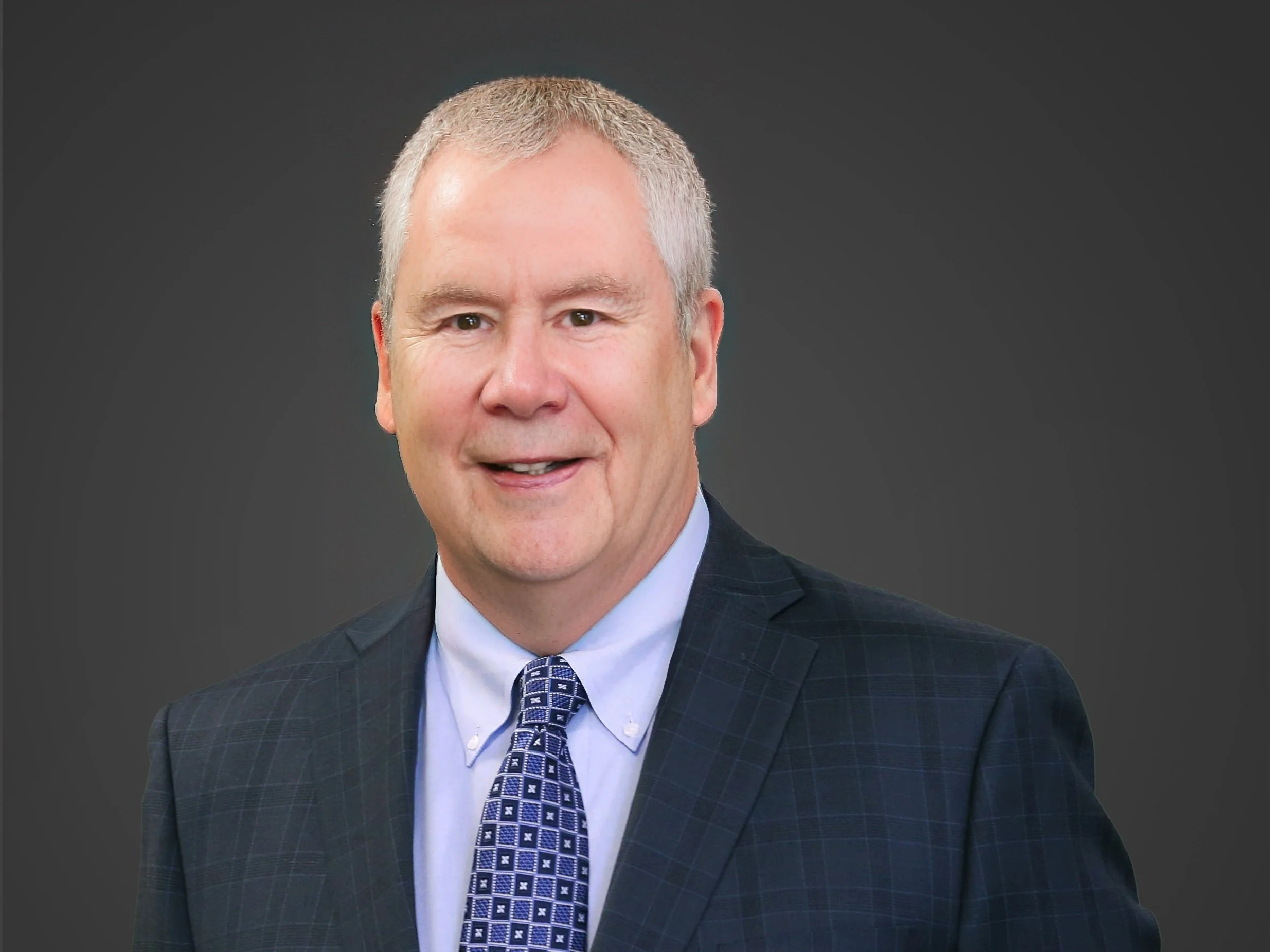 Portrait of a middle-aged man with short gray hair, wearing a dark blue checked suit, a light blue shirt, and a patterned blue tie, against a dark gray background.