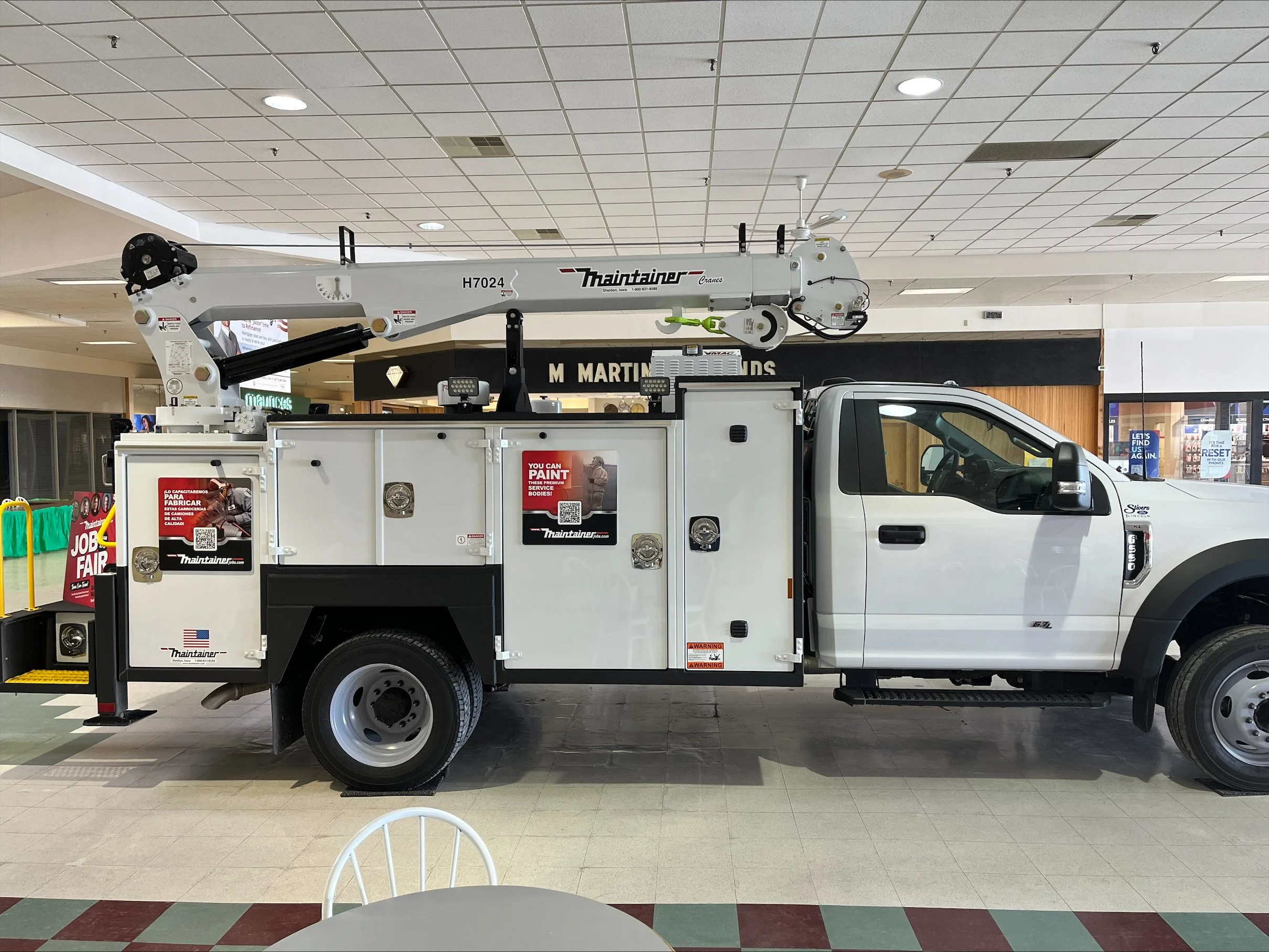 A white utility truck with an articulated boom lift parked inside a shopping mall. The mall has a tiled ceiling and storefronts in the background.