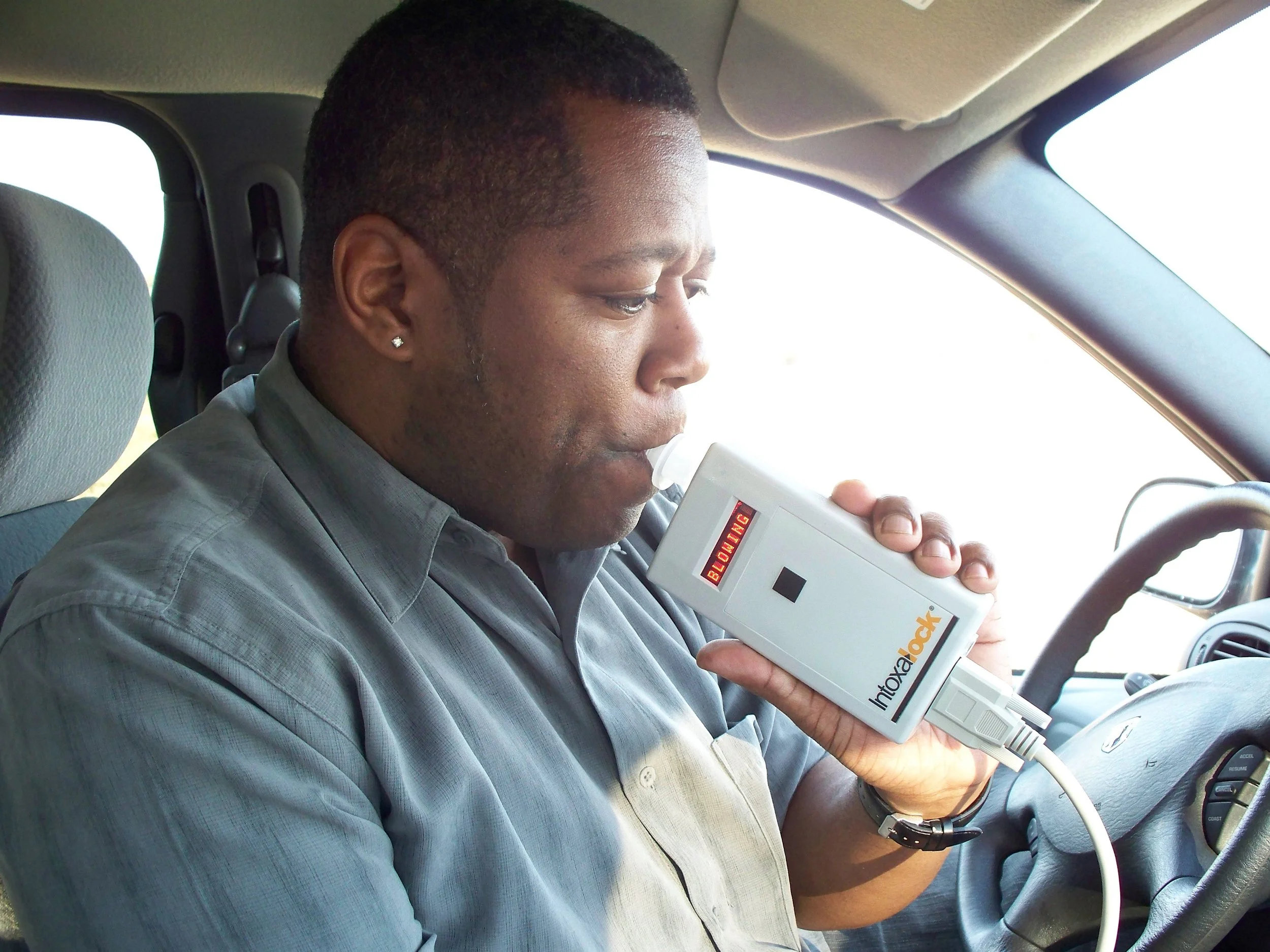 A man with short dark hair, wearing a gray shirt and earrings, is seated in a car and holding a medical testing device close to his lips. The device has a display screen with red and black text and is plugged into the car's dashboard, suggesting a roadside or vehicle-based medical test.