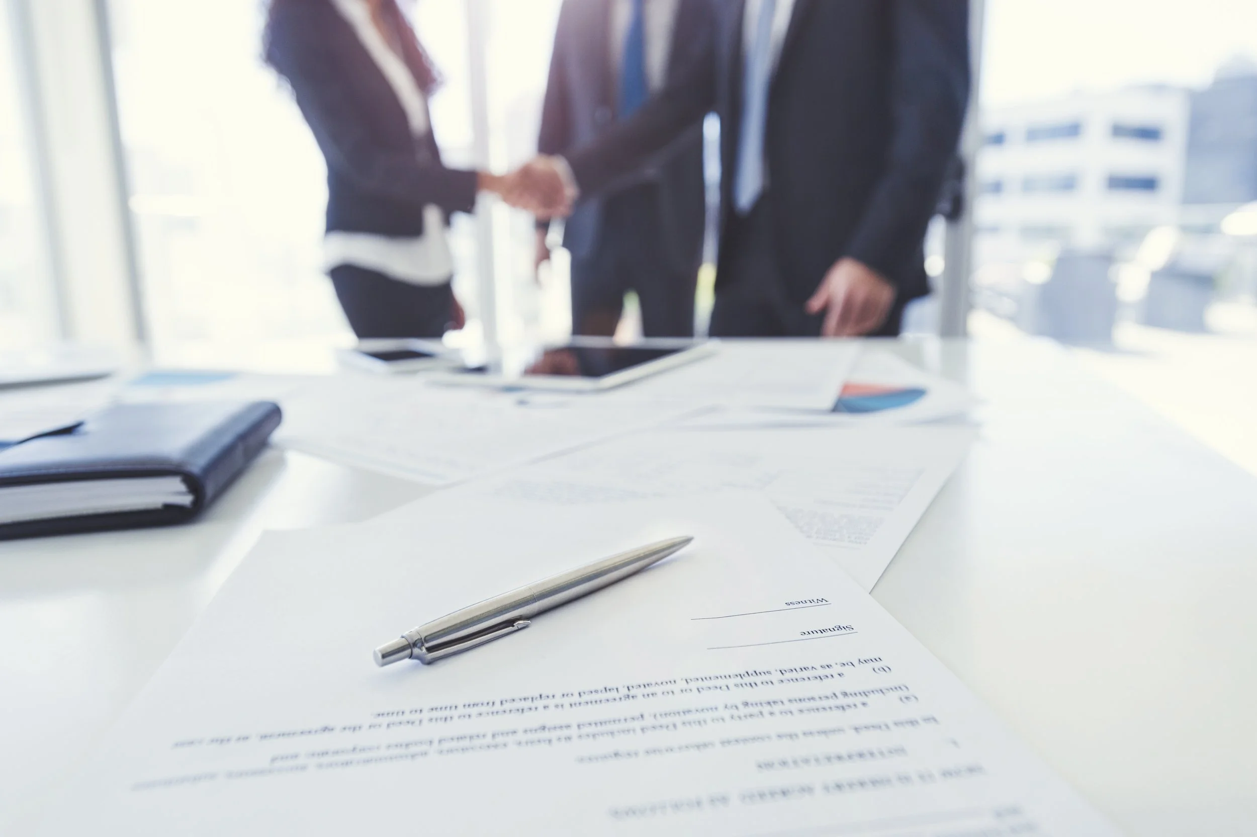 Close-up of a pen and documents on a white table in a bright office, with blurred people shaking hands in the background.