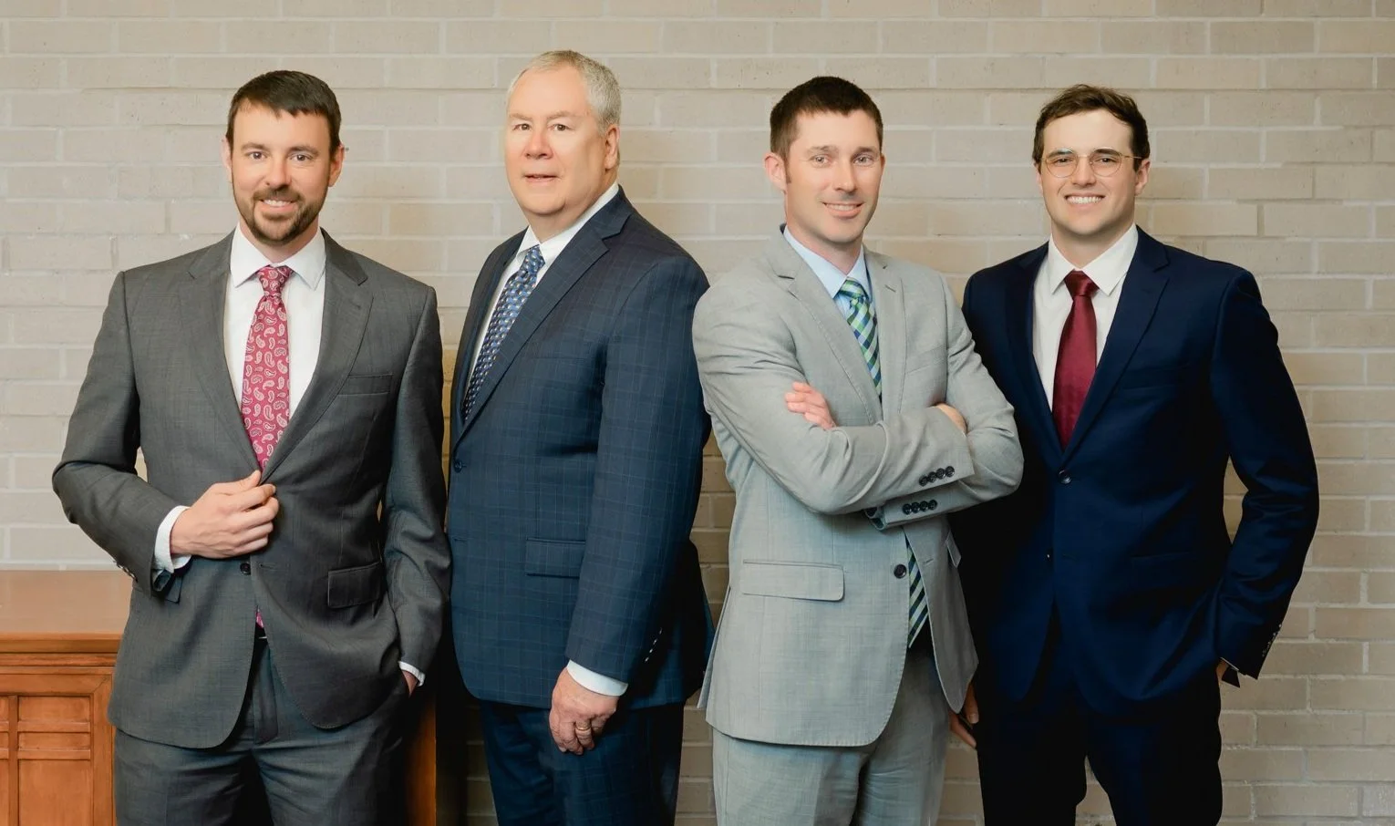 Four men in suits standing side by side in front of a brick wall, looking at the camera.