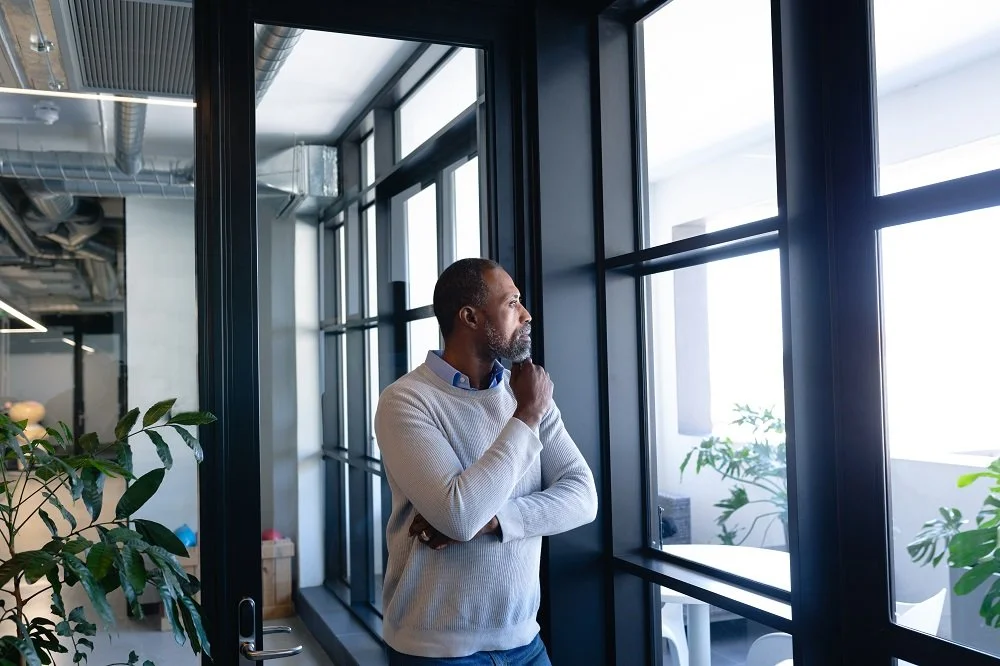A man with a beard and short hair, wearing a light gray sweater, stands next to large windows in a modern office. He is looking outside thoughtfully with one hand on his chin.