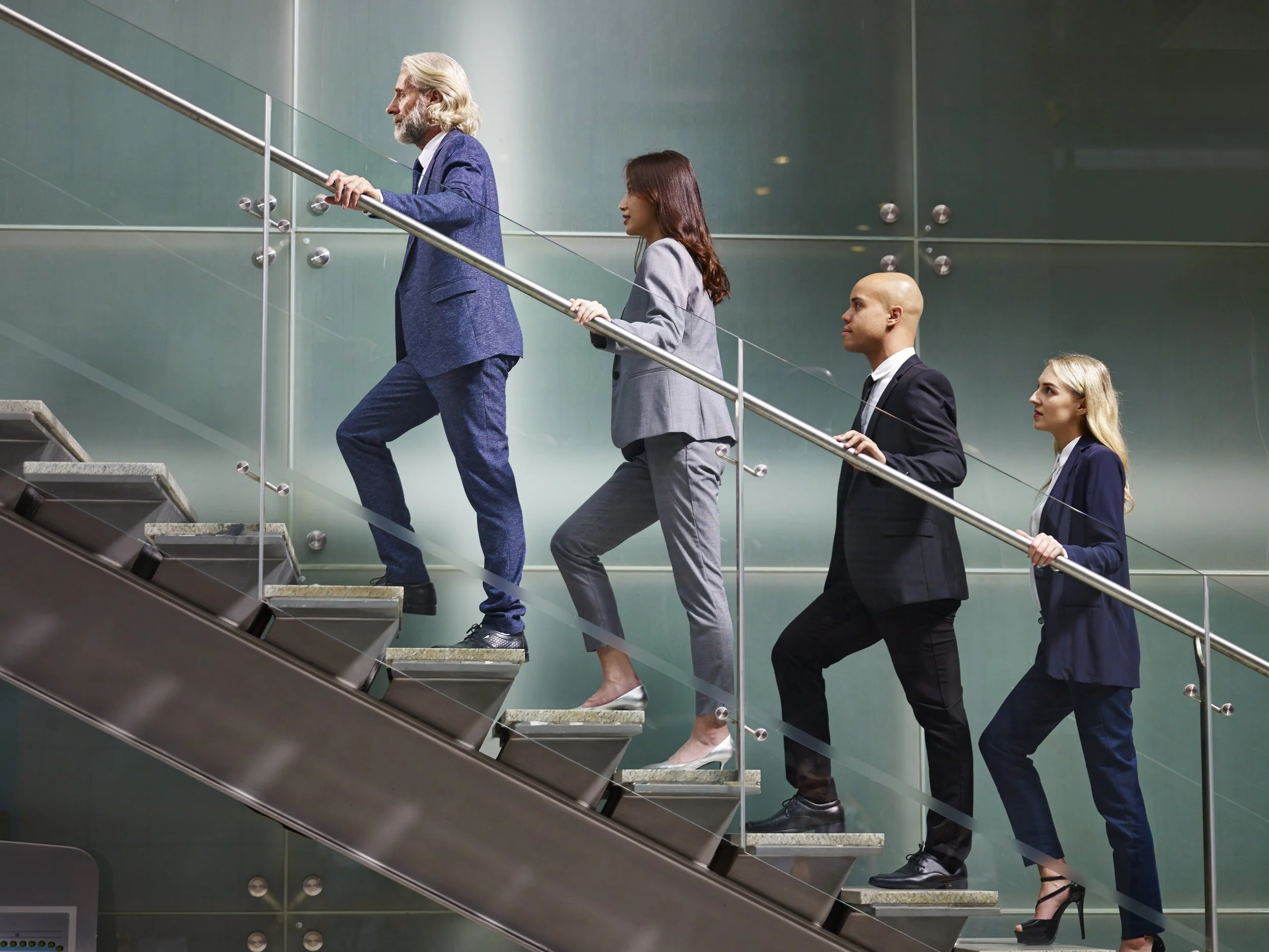 A group of four professionally dressed people, two men in suits and two women in business attire, ascending a staircase in a modern office building, holding onto a metal handrail.