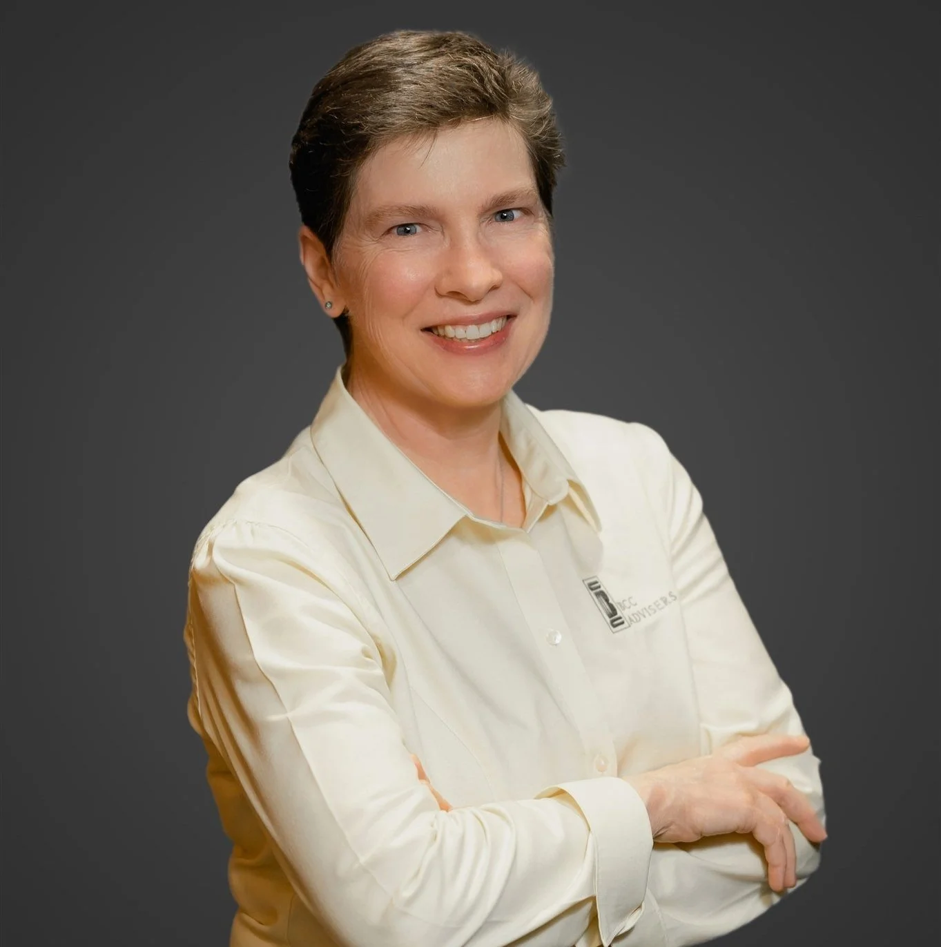 A woman with short brown hair, wearing a cream-colored button-up shirt, smiling with crossed arms against a dark gray background.