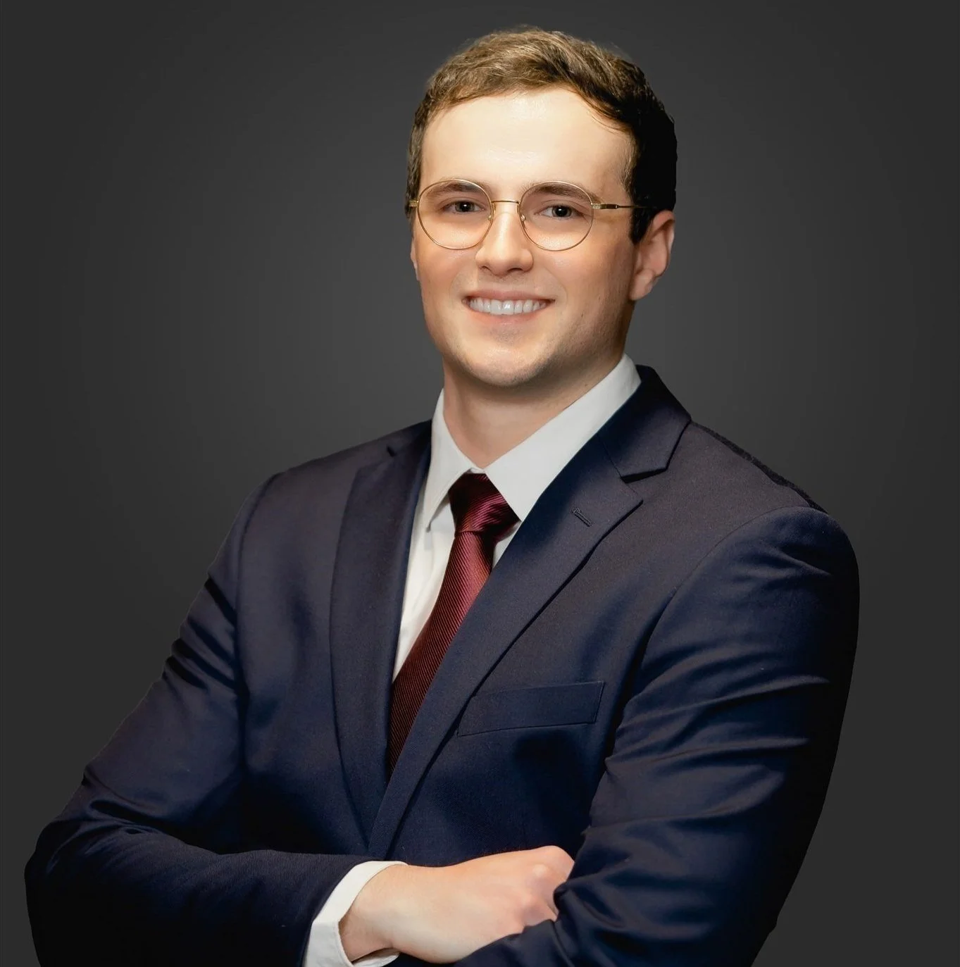 A young man with short brown hair and glasses, wearing a navy suit, white shirt, and maroon tie, smiling confidently with arms crossed against a dark background.