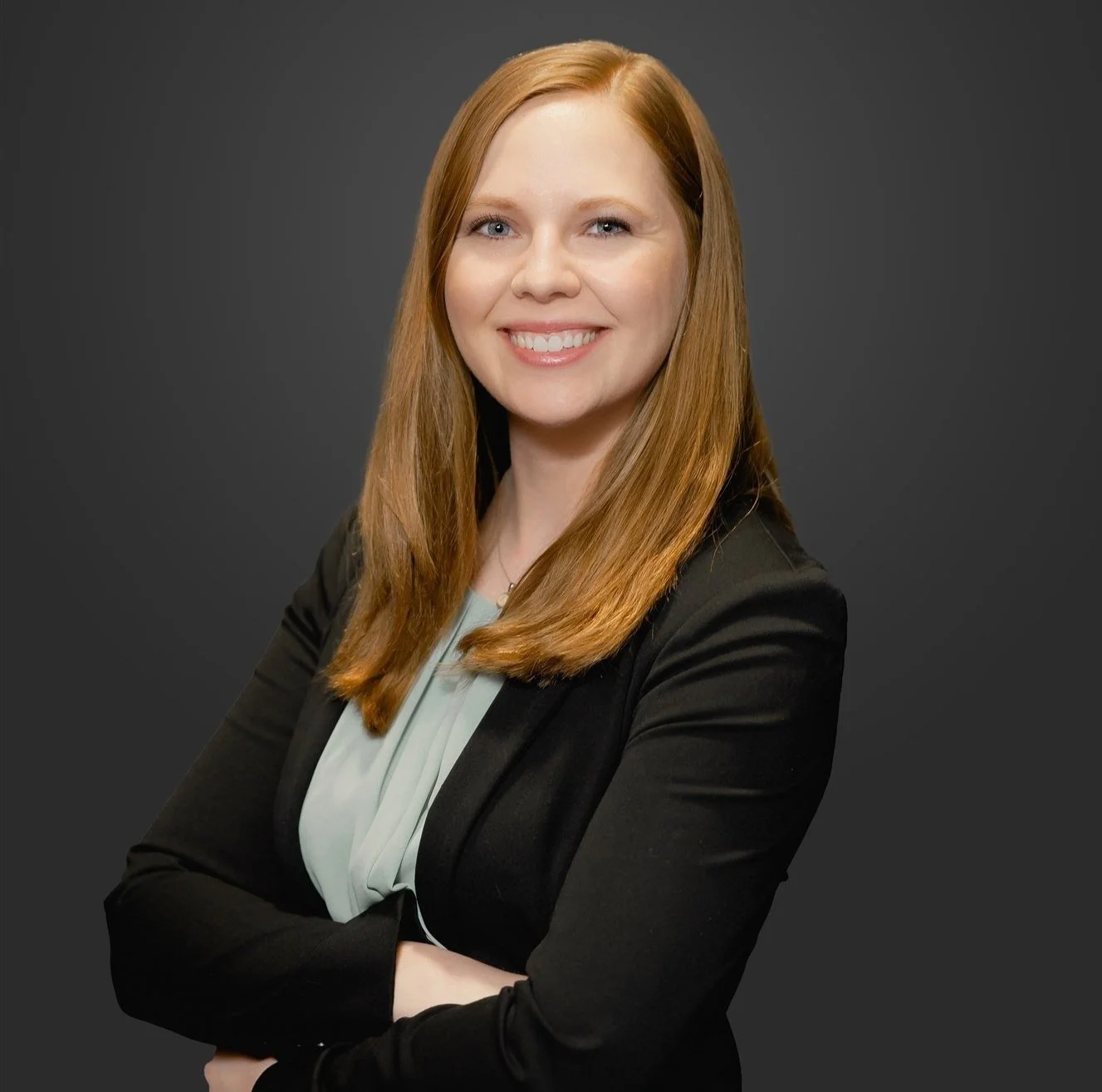 A professional woman with red hair smiling, wearing a black blazer and light-colored blouse, with arms crossed against a dark grey background.