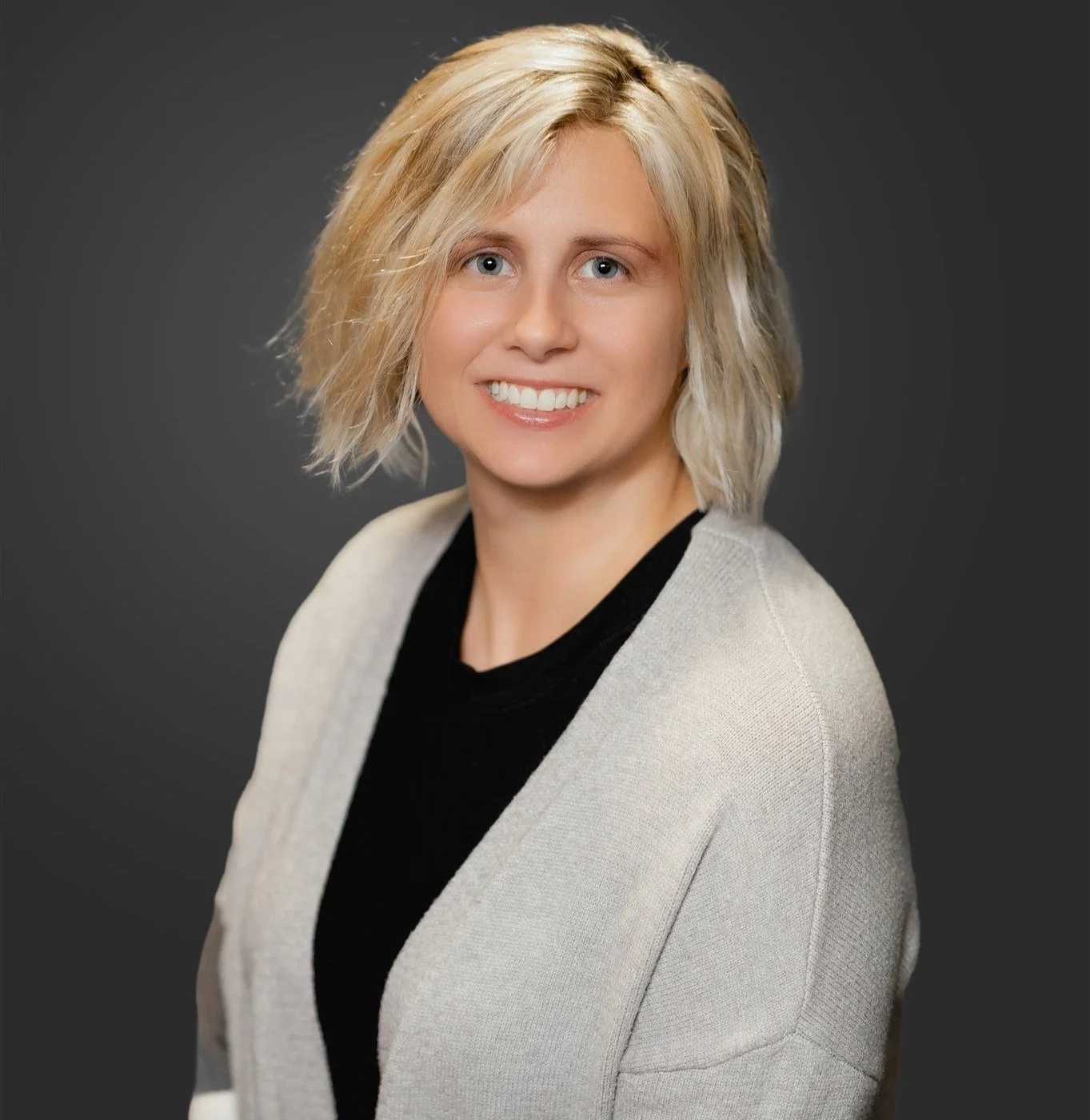 A woman with shoulder-length blonde hair, blue eyes, and a bright smile, dressed in a light gray blazer over a black top, posing against a dark gray background.