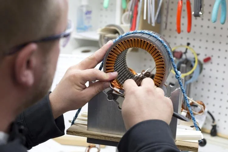 A person working on an electric motor, examining its copper coils and structure, with tools hanging on a pegboard in the background.