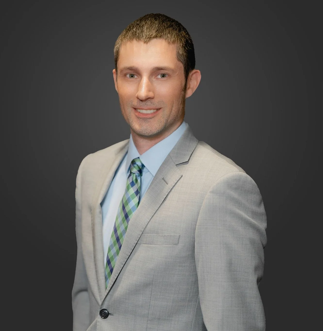 A portrait of a man with short brown hair, wearing a light gray suit, light blue shirt, and a plaid tie, smiling against a dark background.
