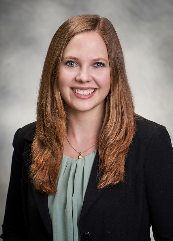 Portrait of a young woman with long red hair smiling, wearing a black blazer over a light green blouse, with a small necklace, against a neutral gray background.