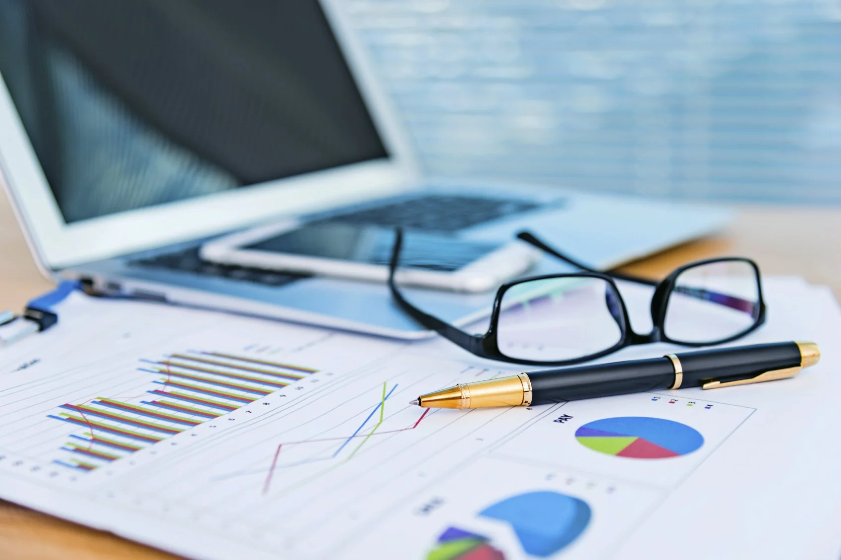 Work desk with financial charts, a pen, glasses, a smartphone, and a laptop, suggesting a business or investment analysis setting.