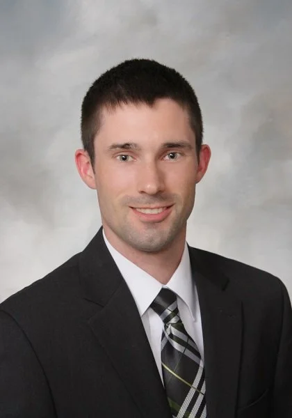 Portrait of a young man in a black suit, white shirt, and plaid tie, smiling against a neutral background.