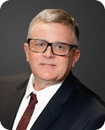 Portrait of a man with short light-colored hair, glasses, wearing a dark suit, white shirt, and maroon tie against a gray background.