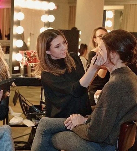 Makeup artist applying makeup to woman in a backstage setting with bright vanity lights.
