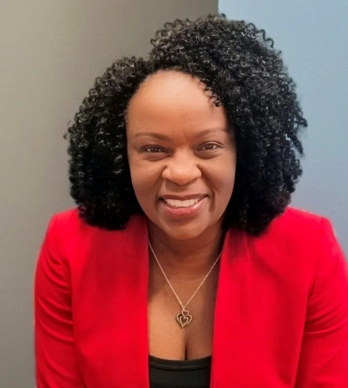 A smiling woman with curly black hair, wearing a red blazer, black top, and a silver necklace with a heart pendant, standing against a blue-gray wall.
