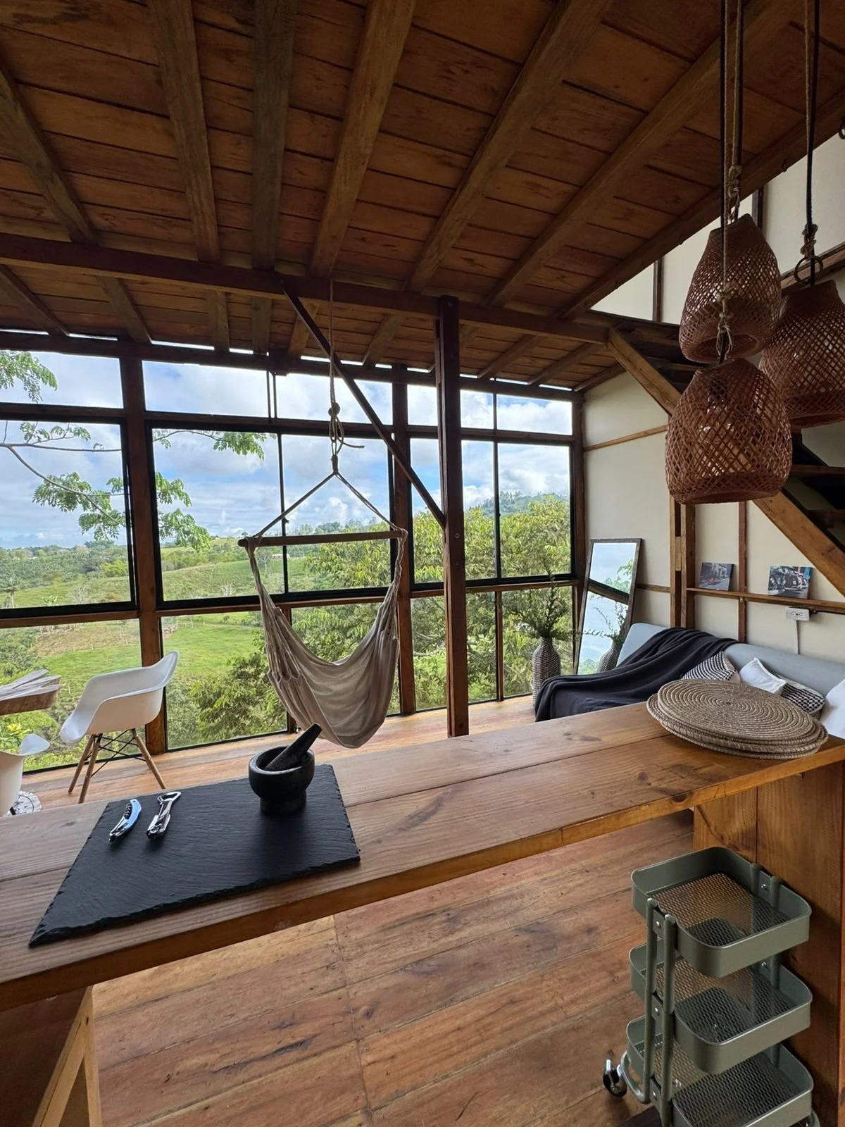 Interior de una casa con paredes de cristal y techo de madera, con vistas a un paisaje verde. Hay una silla blanca, un columpio de tela, una mesa con utensilios y platos de mimbre colgados del techo.