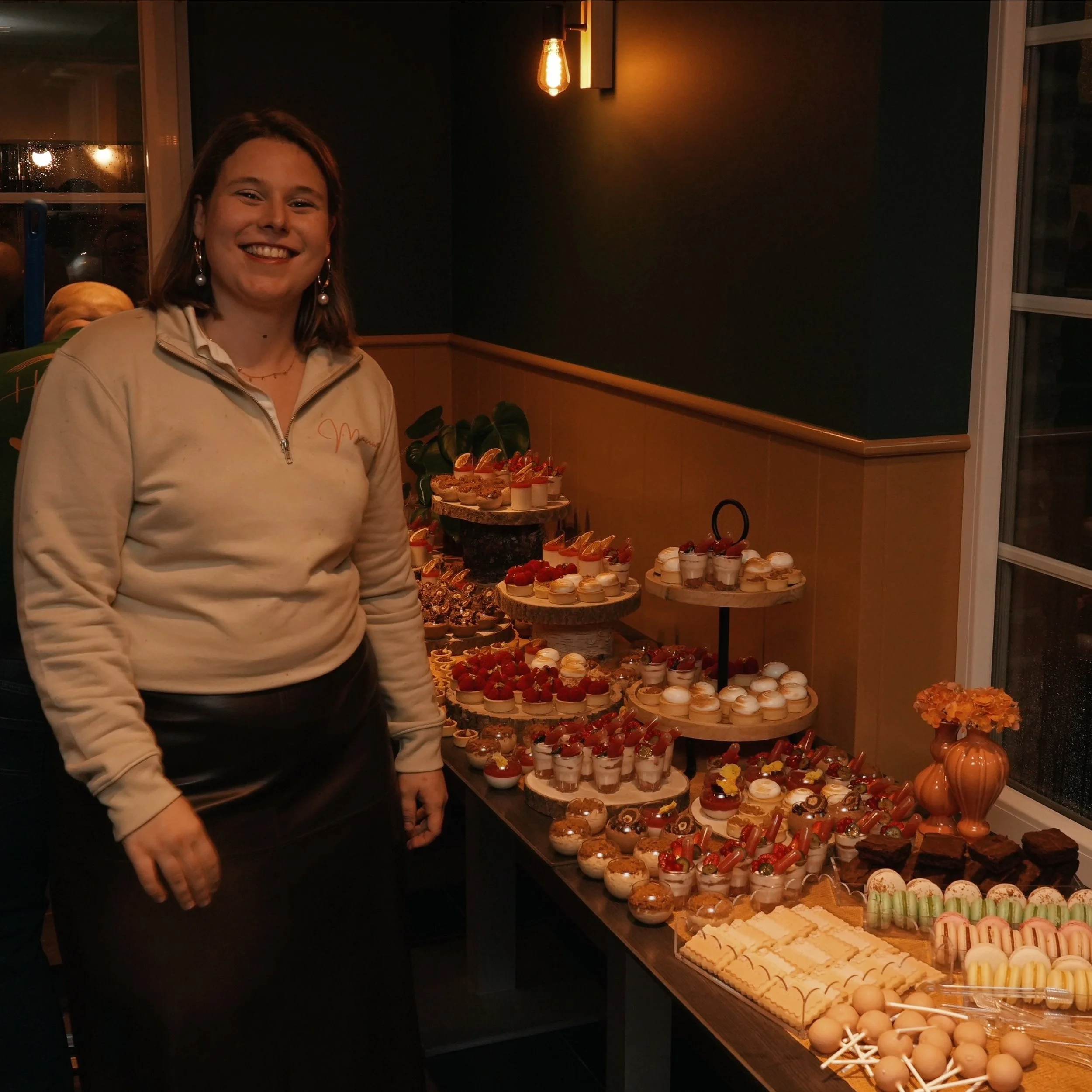 A woman with shoulder-length hair and earrings smiling at a dessert table filled with various miniature desserts and cupcakes in a dimly lit room with warm lighting.