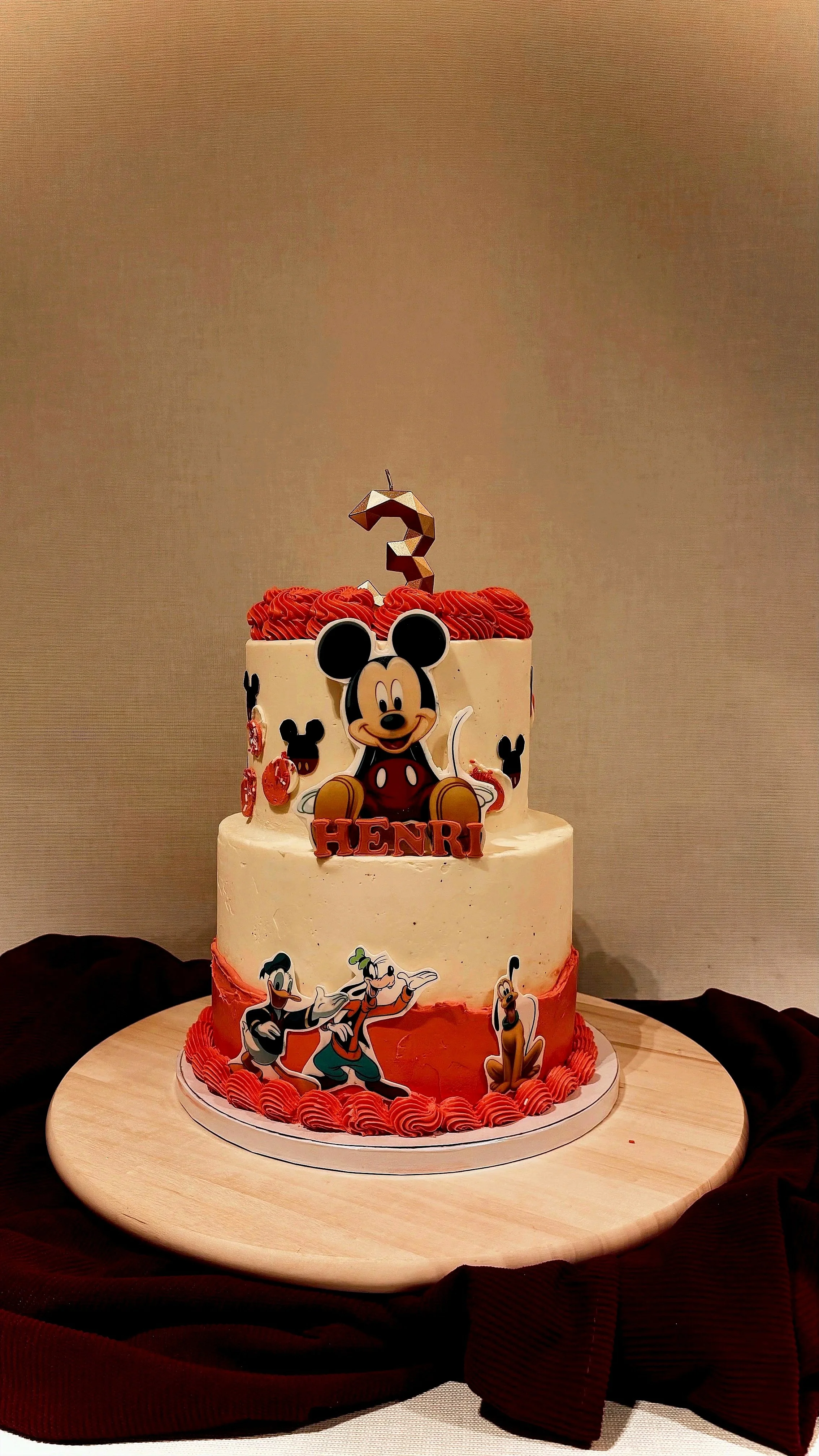 A two-tier birthday cake decorated with Mickey Mouse and Disney character images. The cake features a Mickey Mouse topper, a number 3 candle, and the name 'Henri' written on it, set on a wooden cake stand with a dark red cloth underneath.